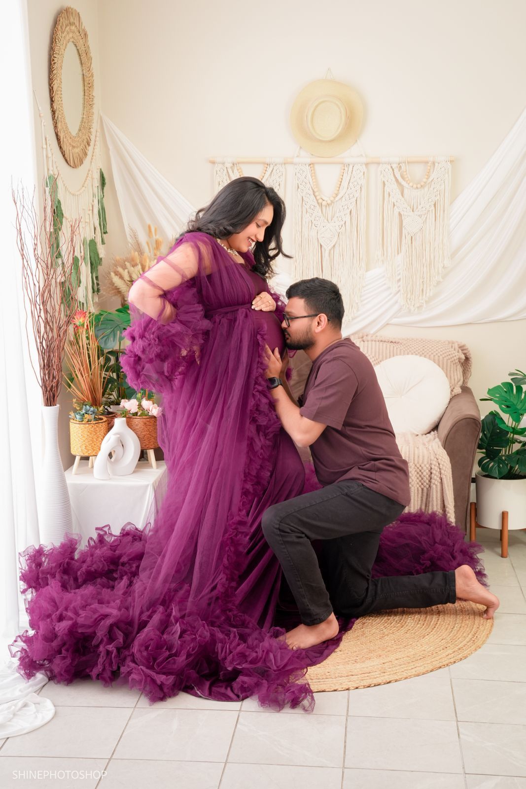Couple in boho studio, woman in purple tulle maternity gown, man kneeling kissing her belly, warm decor backdrop.