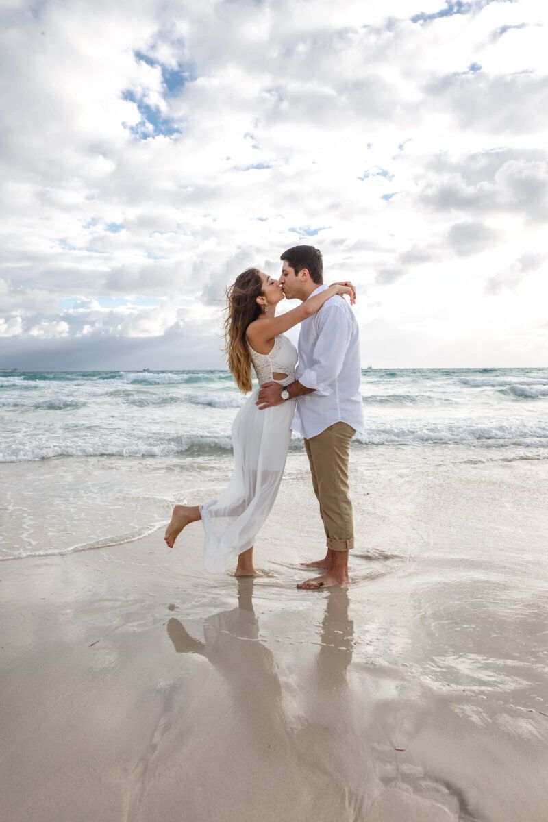 A couple shares tender moments on a bright sandy beach with ocean waves in the background.