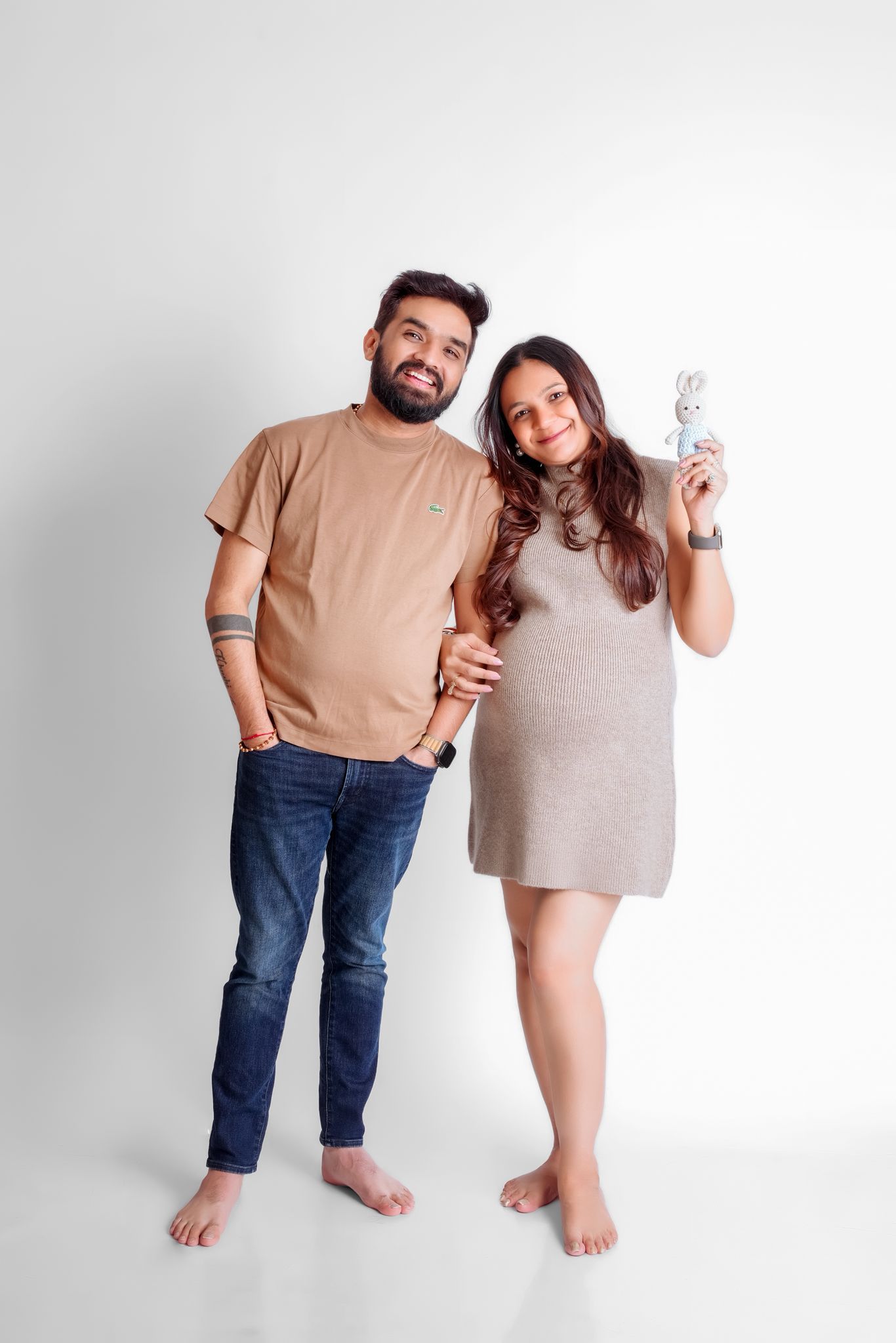 Casual maternity portrait with couple in matching neutral tones against white background.