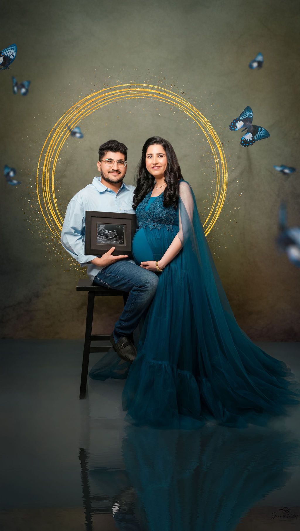 Couple poses with ultrasound photo against golden circle backdrop with blue butterflies.