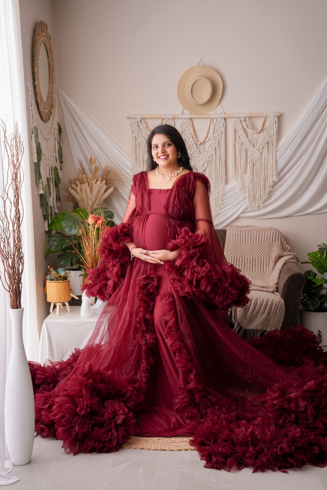 Pregnant woman in elegant burgundy ruffled tulle maternity gown posing in boho studio setting.