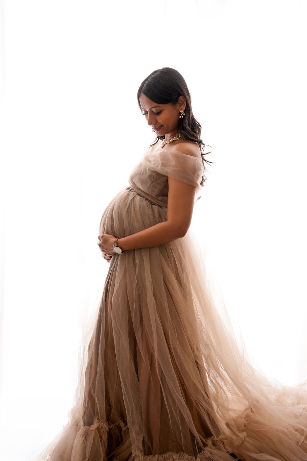 Pregnant woman in elegant beige tulle gown cradling baby bump, posing gracefully against white background.