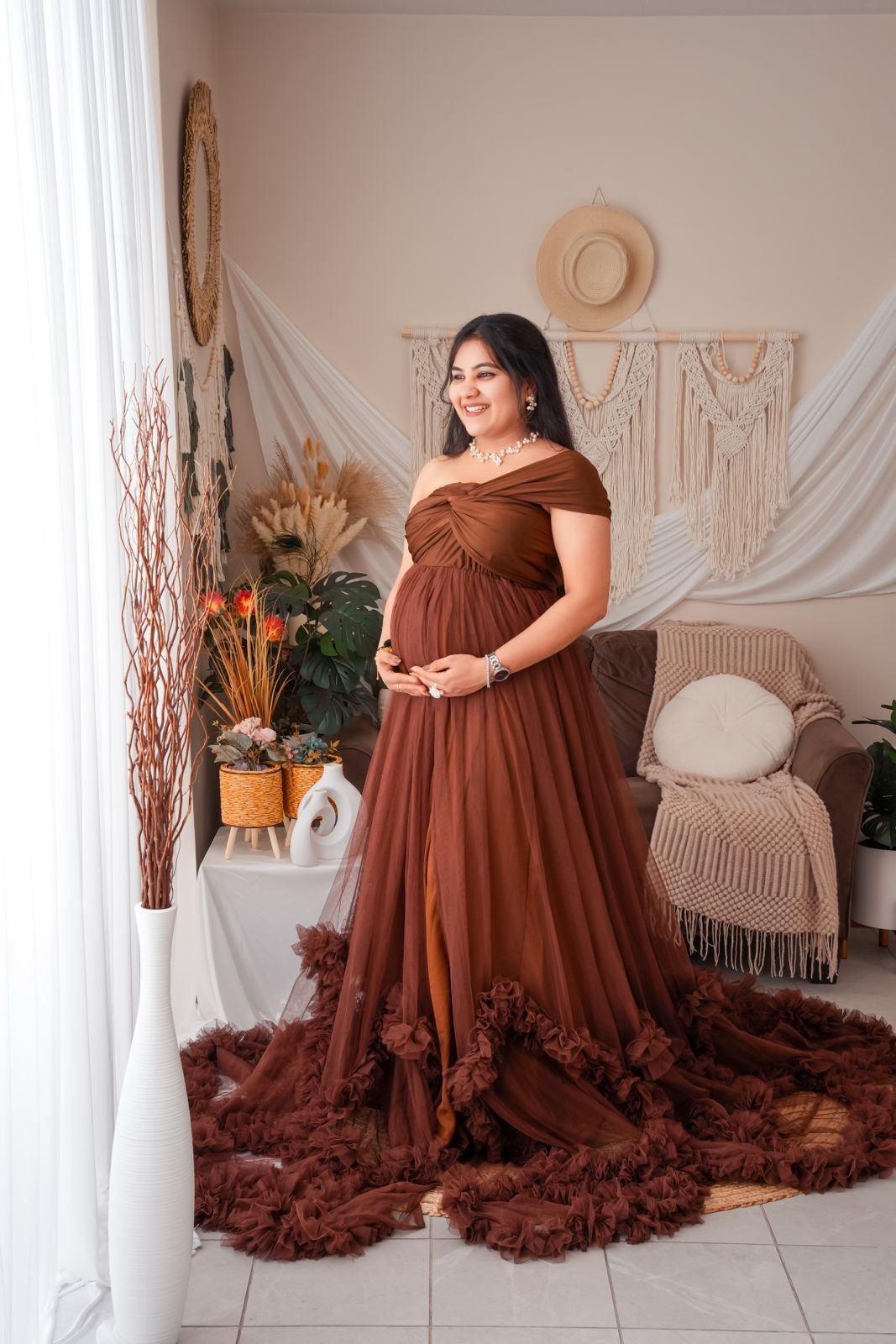 Pregnant woman in elegant rust brown off-shoulder tulle maternity gown posing in boho-styled studio with macrame decor.