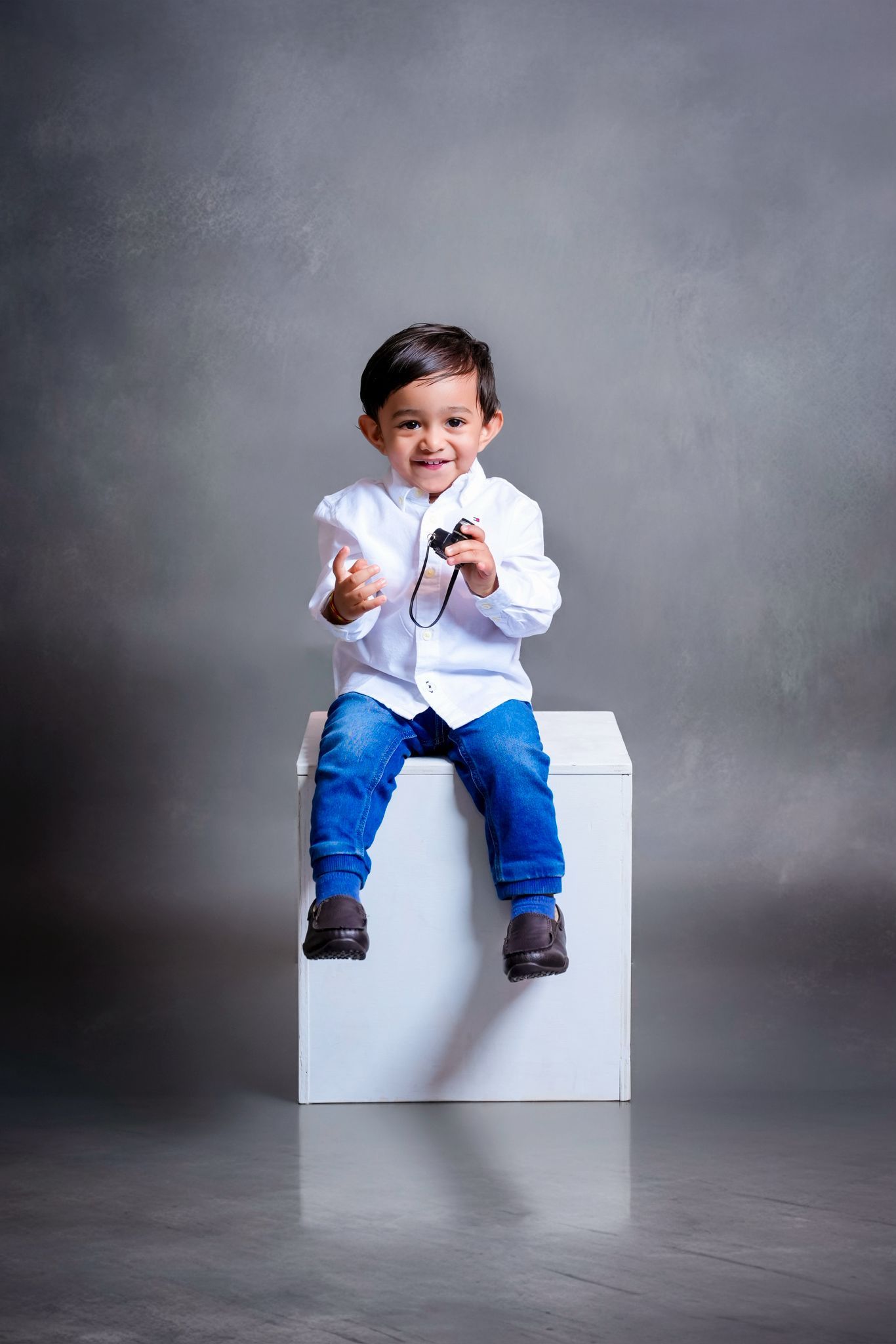 A toddler in blue jeans and white shirt sits on a white cube against a gray background in a photo series.