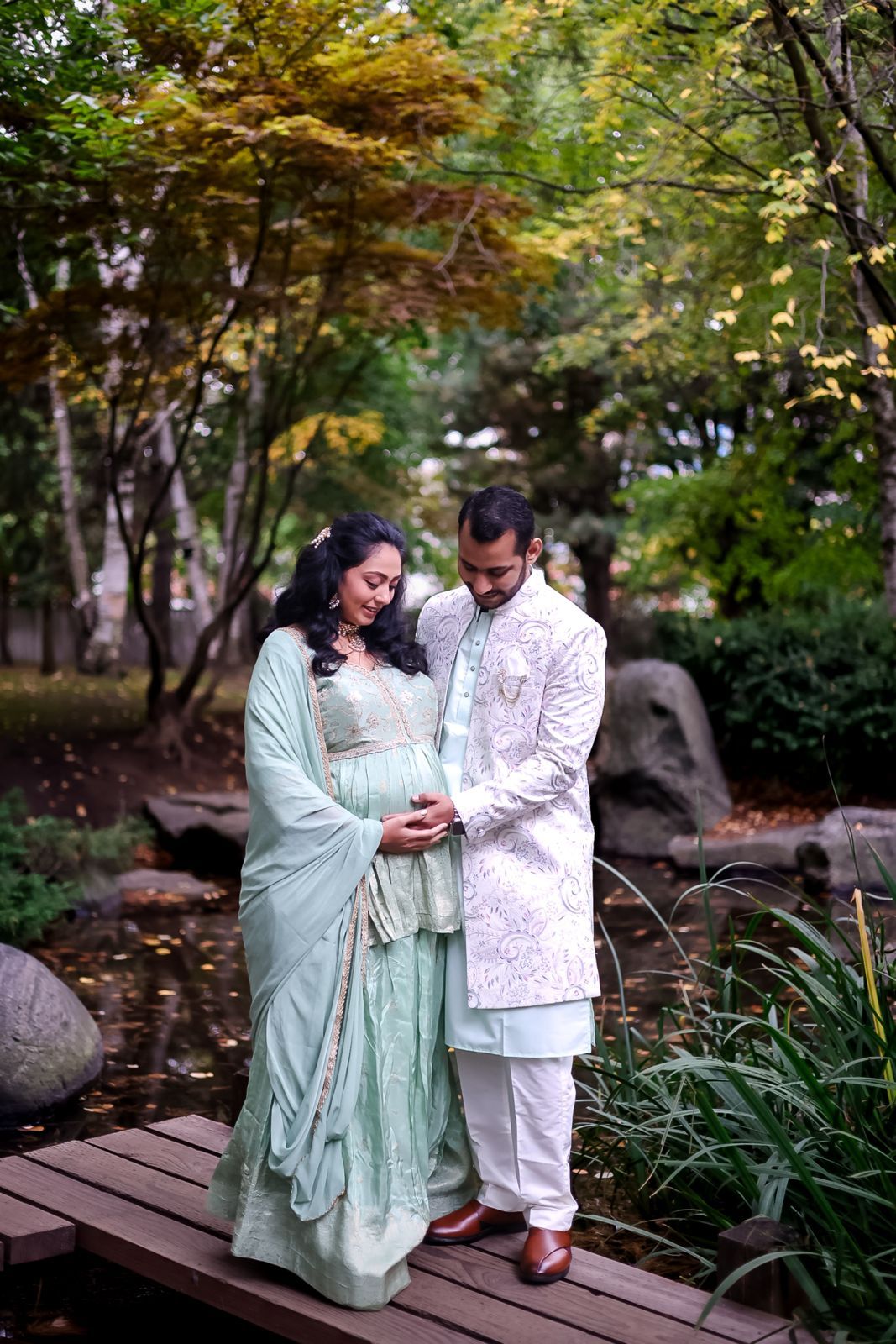A couple in elegant mint green and white traditional attire poses on a wooden bridge in a garden.