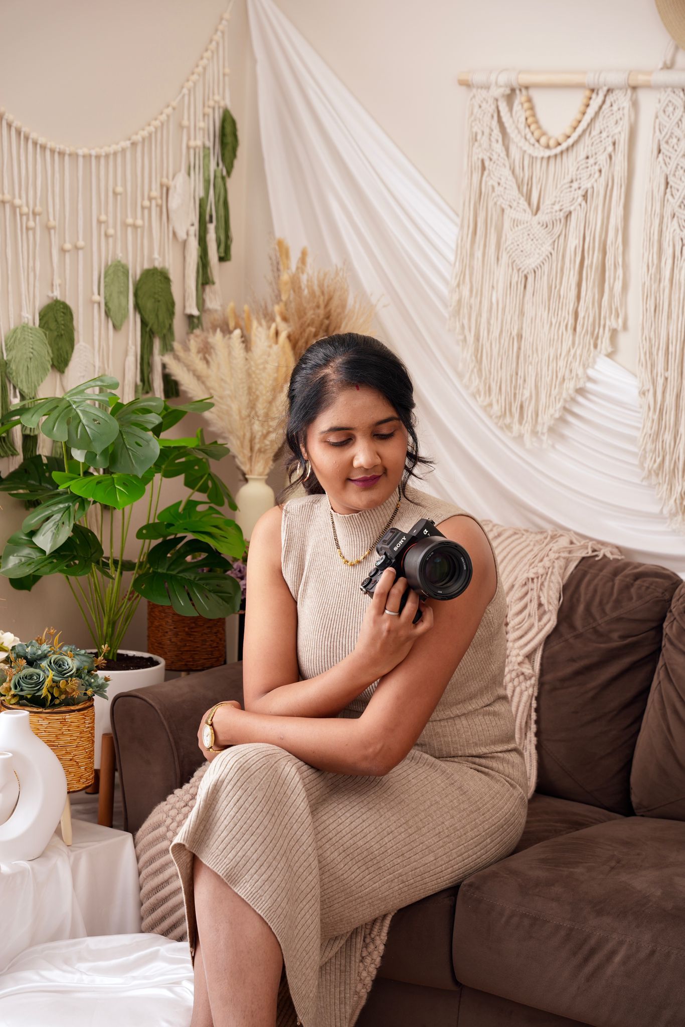 A photographer poses with a camera on a brown couch with macrame wall hangings and plants in the background.