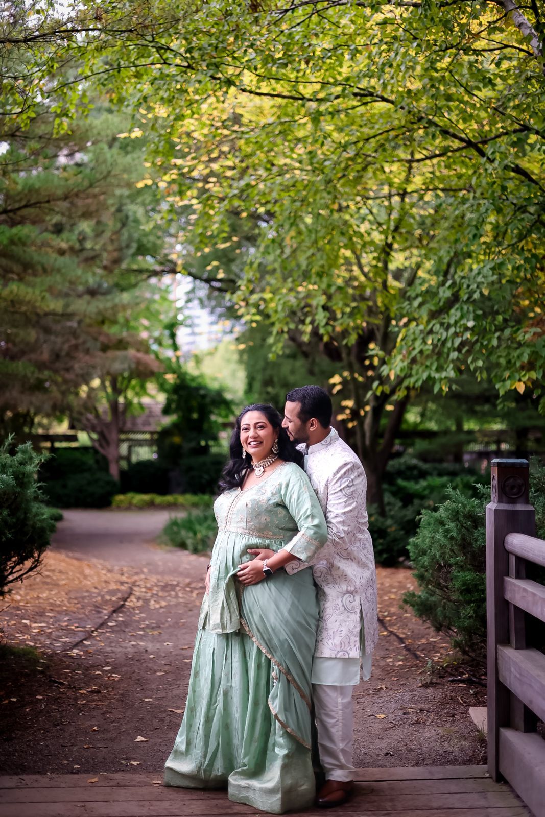 A couple in mint green traditional attire poses on a wooden pathway surrounded by trees.