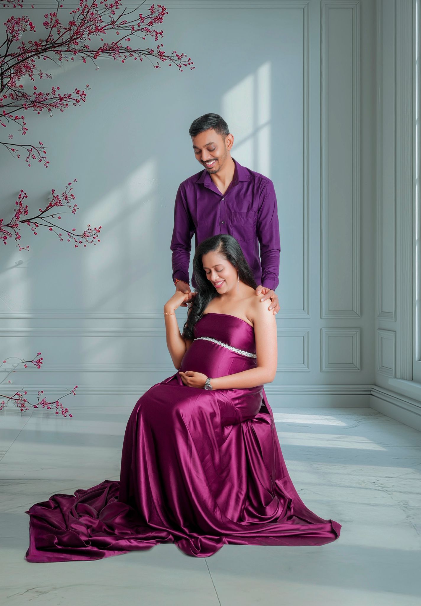 A couple in purple attire poses together in a romantic maternity photoshoot against a light blue wall with cherry blossoms.
