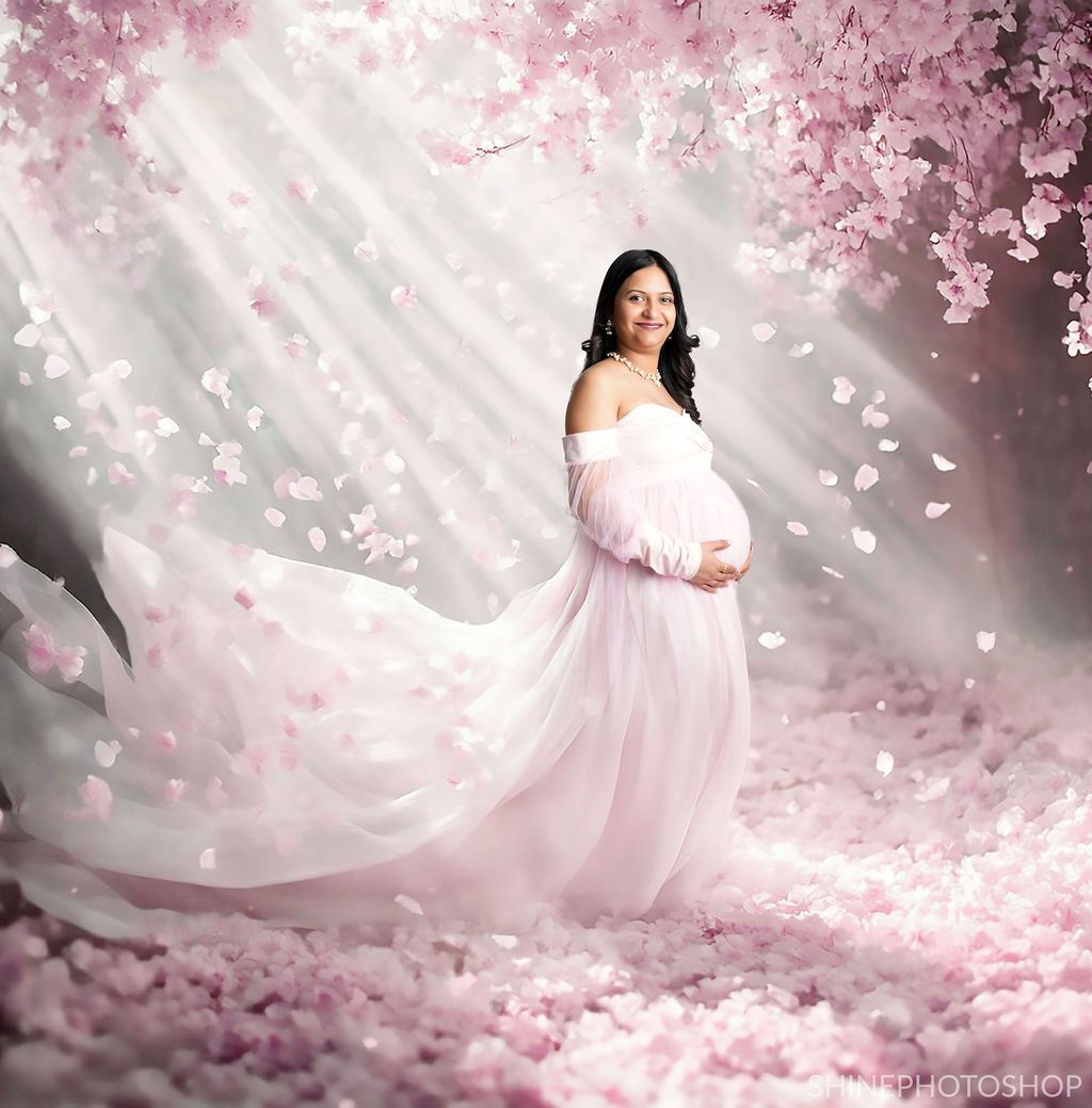 Expectant parent in flowing pink gown poses amid cherry blossoms and dreamy light rays.