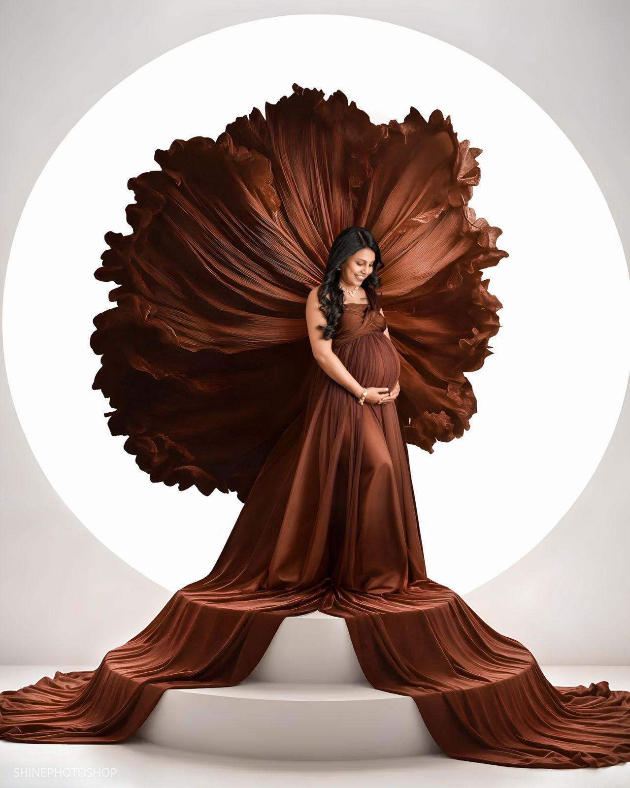 Pregnant woman in flowing rust-brown satin maternity gown with dramatic billowing fabric against white circular backdrop.