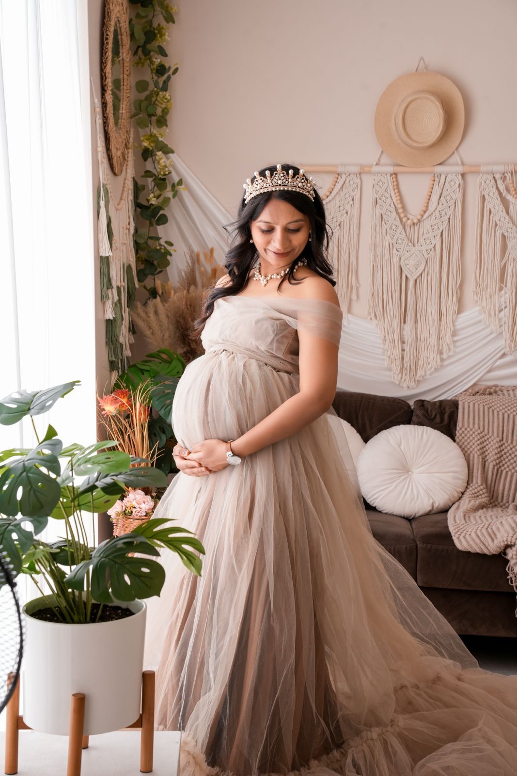 Pregnant woman in elegant taupe tulle gown and tiara poses in boho-styled room with plants and macrame decor.