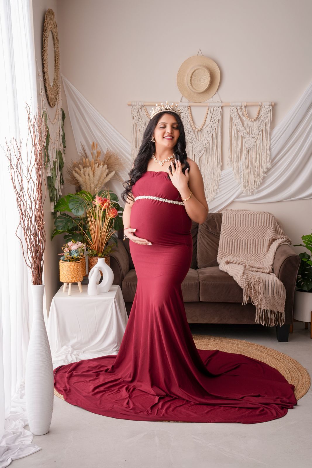 Pregnant woman in elegant burgundy mermaid gown posing in boho studio with macrame decor and plants.