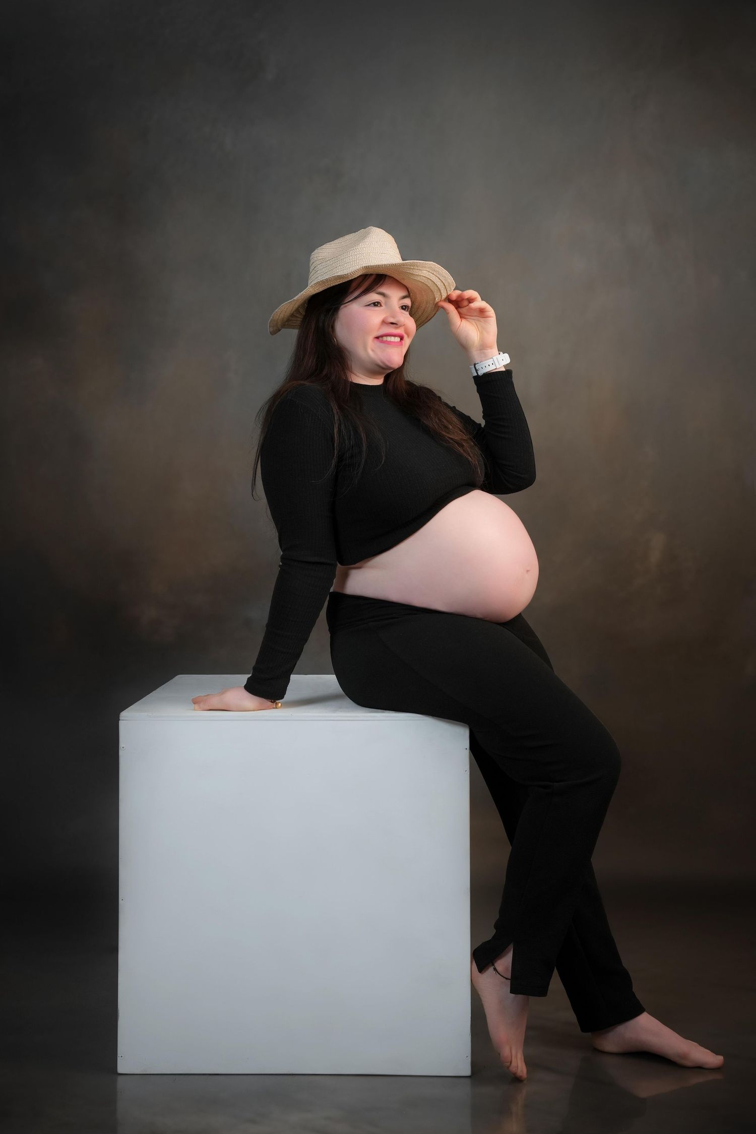 Pregnant woman poses on white cube during artistic studio maternity session.