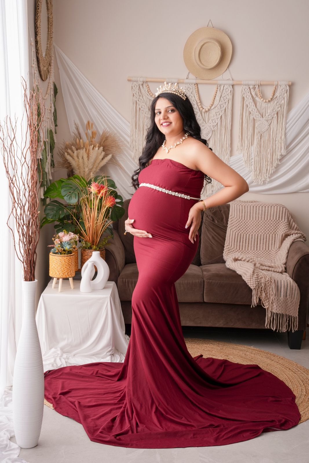 Pregnant woman in elegant burgundy mermaid gown with pearl belt and crown poses in boho-styled studio.
