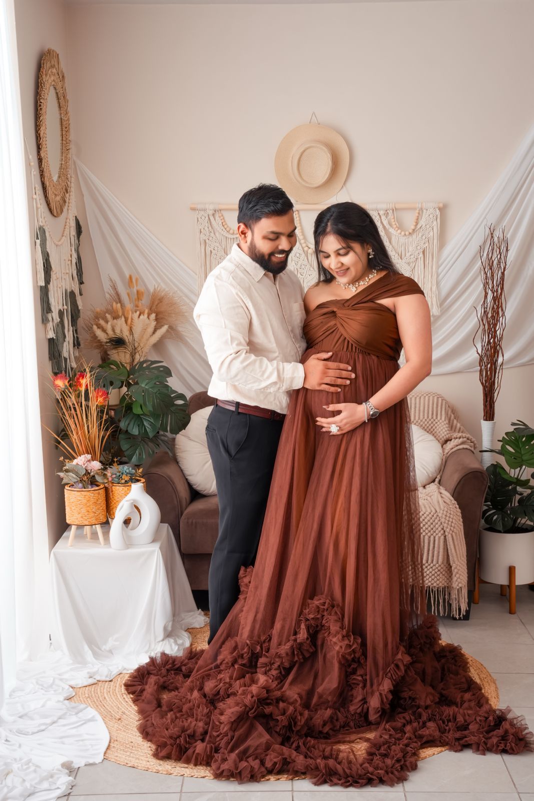 Couple in boho studio, pregnant woman in brown tulle gown, partner in white shirt, maternity photoshoot.