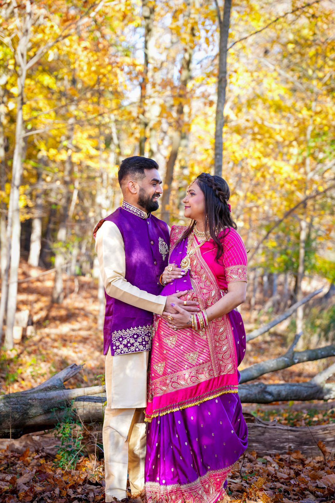 A couple in purple and pink Indian outfits poses playfully among autumn leaves.