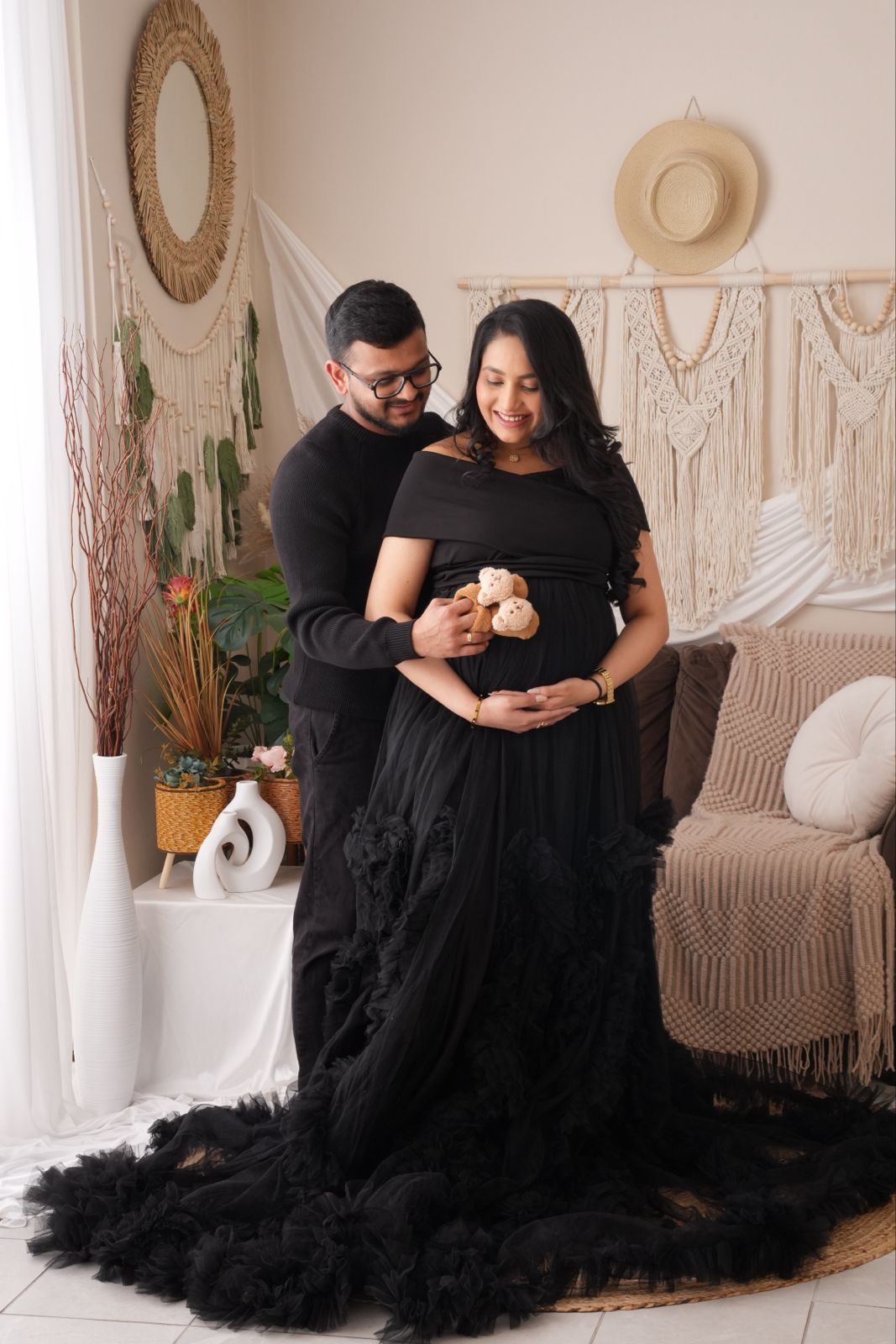 Couple in black outfits during maternity photoshoot in boho studio, pregnant woman in ruffled gown holding teddy bear.