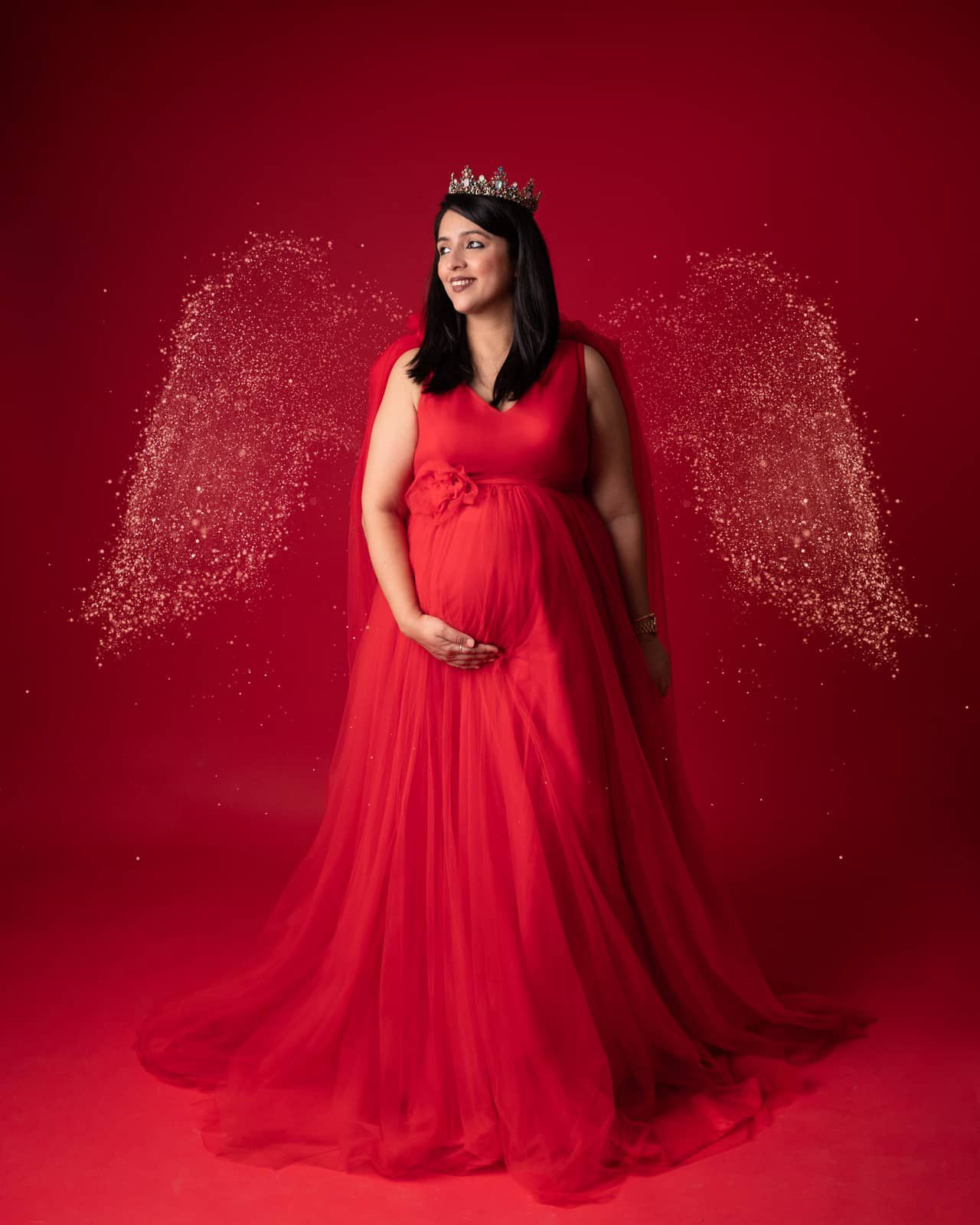 Stunning red tulle gown with glitter effects in maternity photos against a deep crimson backdrop.