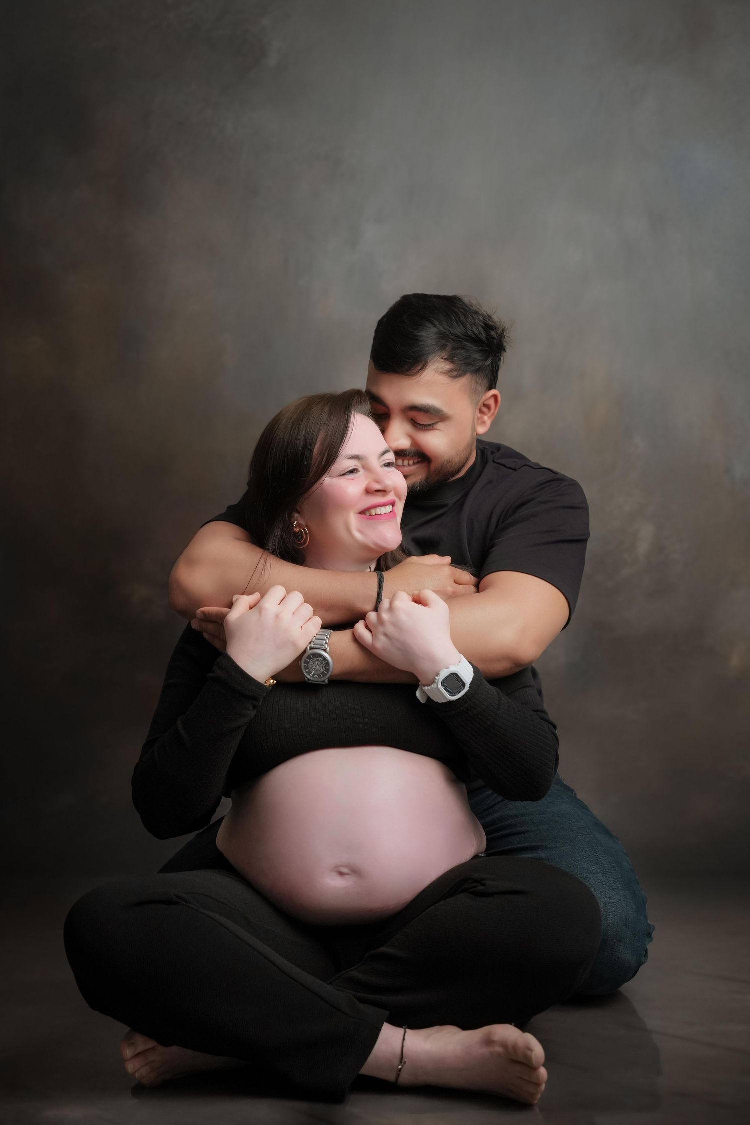 Intimate maternity photography session with couple in black against moody backdrop.