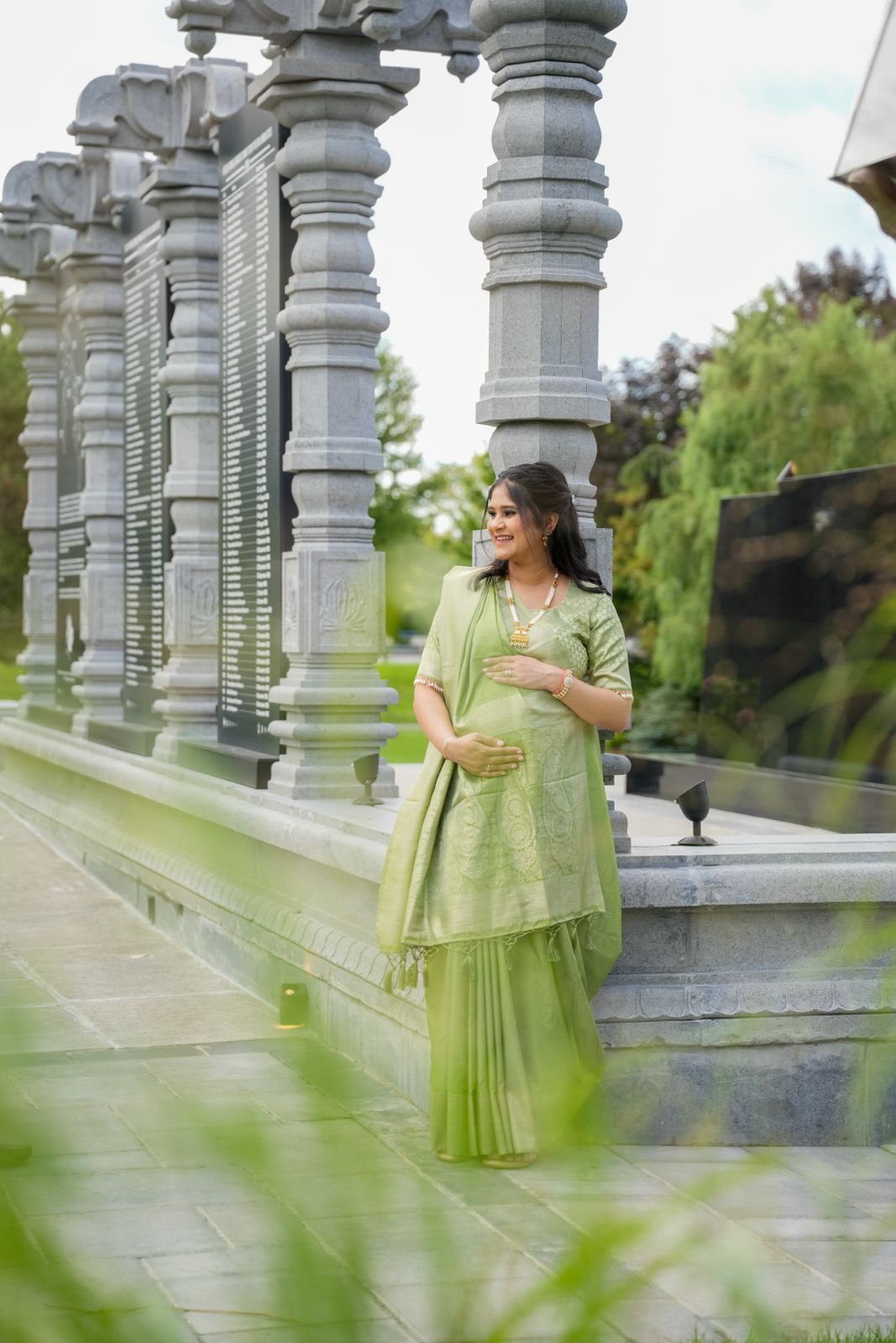 A person in a mint green traditional outfit poses by ornate stone pillars in a garden setting.