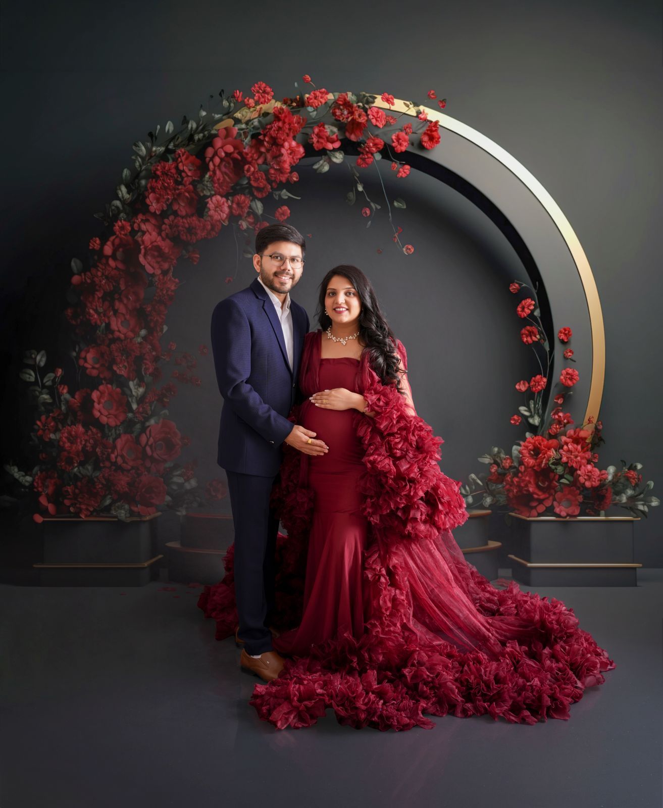 Couple poses for maternity photo in front of red floral arch, woman in red gown cradling baby bump, man in navy suit.