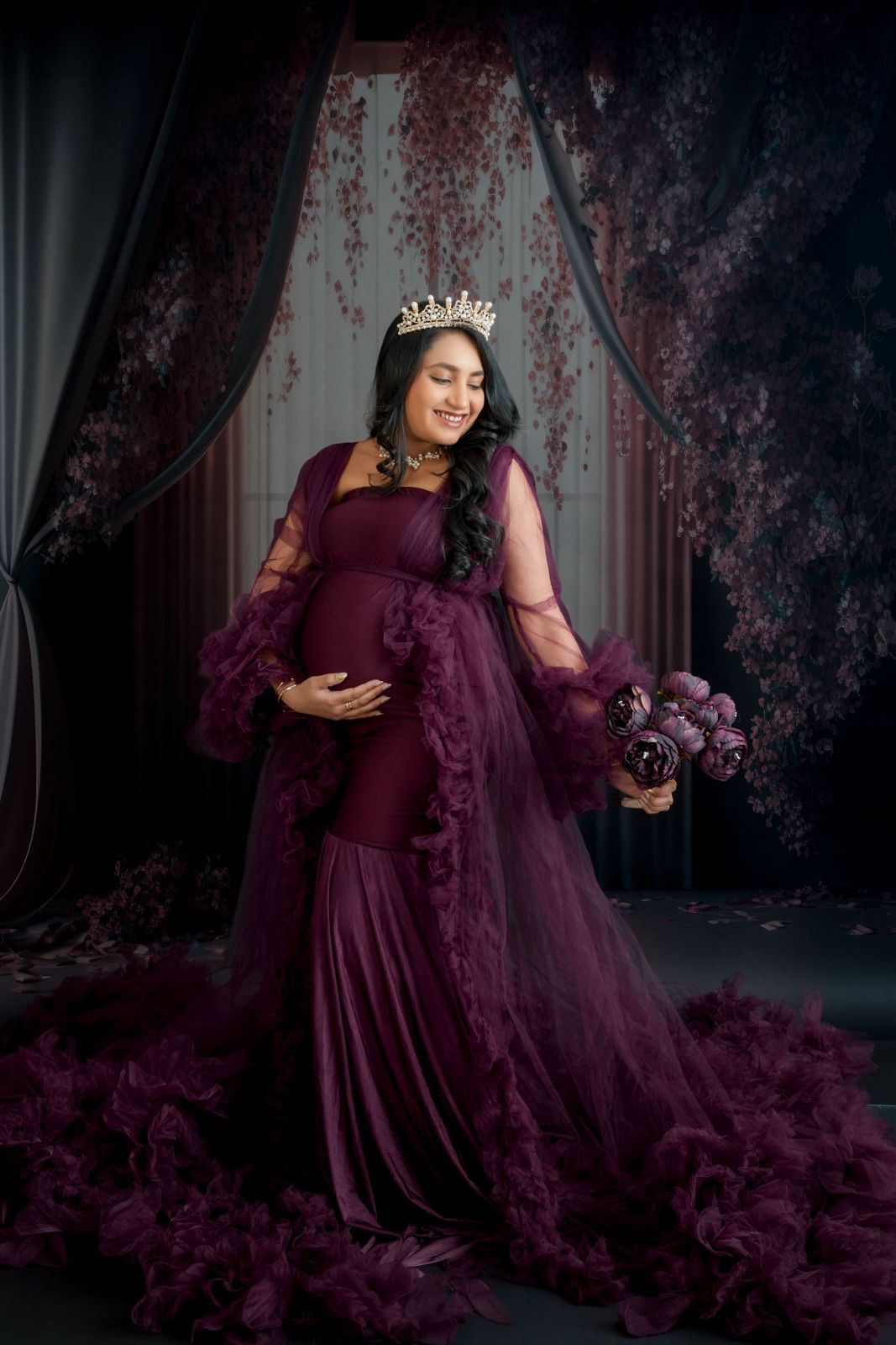 Pregnant woman in elegant burgundy gown with crown and floral bouquet poses for maternity portrait.