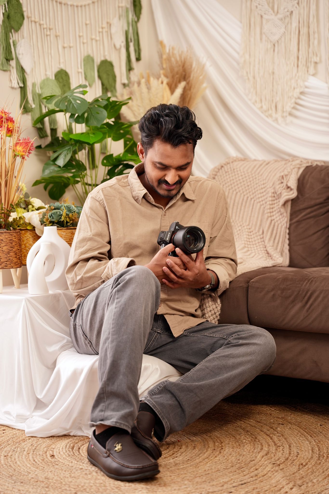 Photographer sitting on floor reviewing photos on digital camera in cozy boho room with plants and macrame decor.