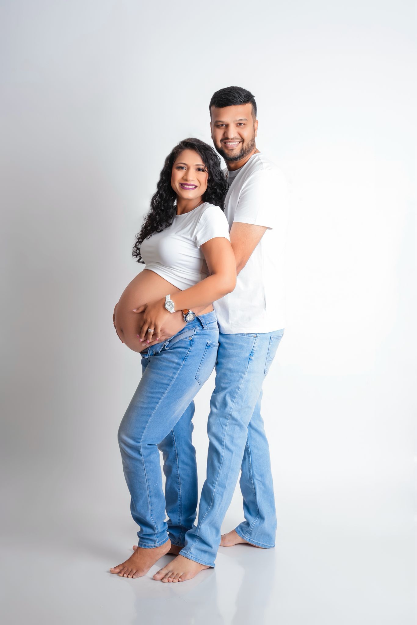 Casual maternity pose with couple in denim and white against plain background.