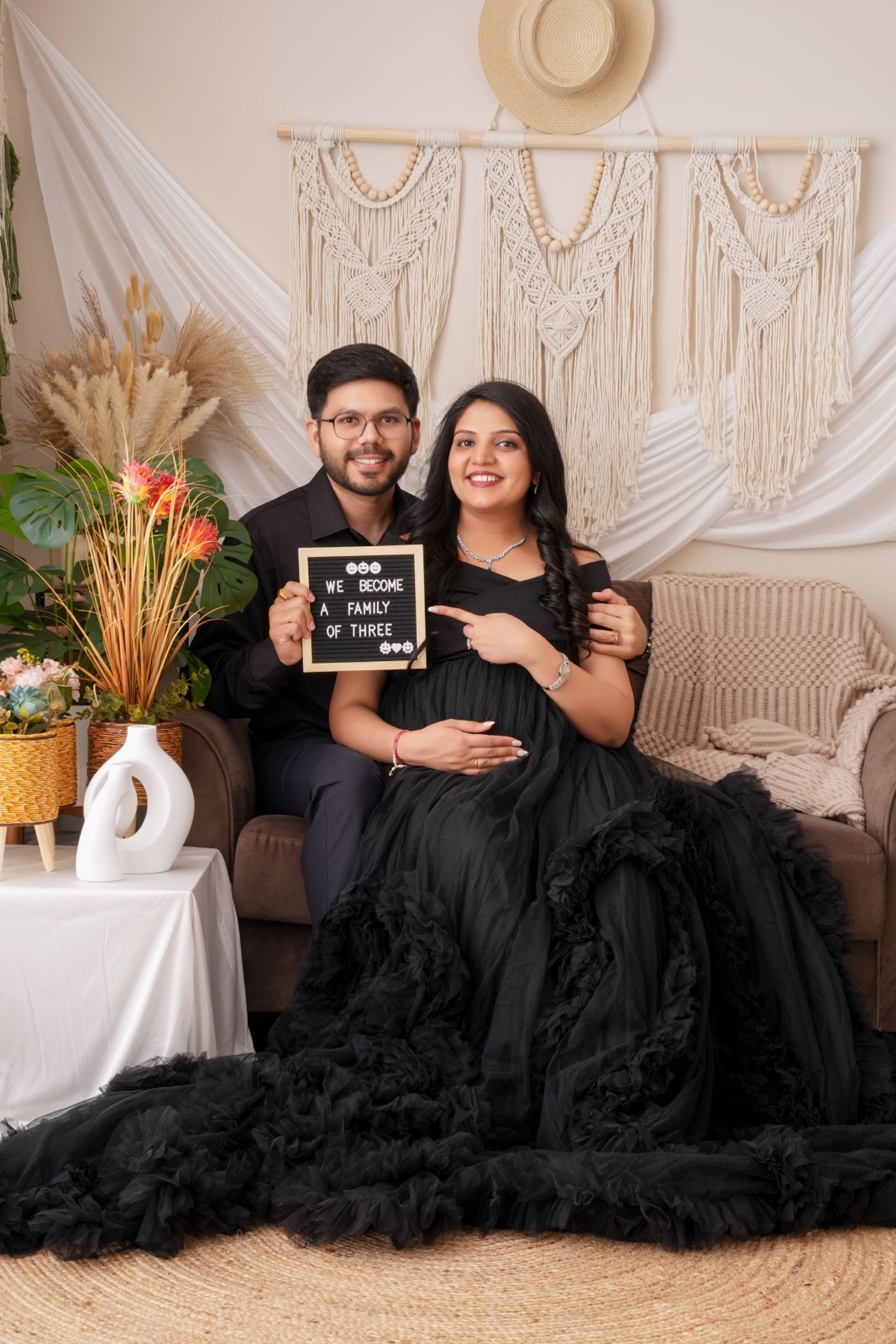 Couple in black outfits holding a family of three sign, maternity photoshoot with boho decor backdrop.