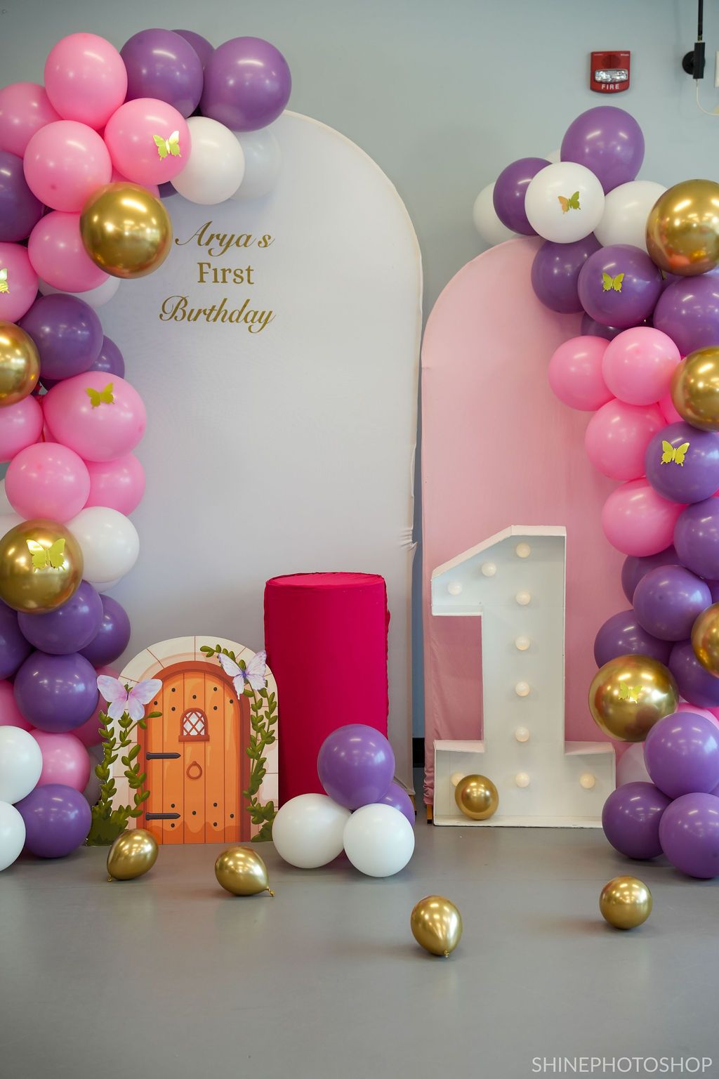 First birthday party backdrop with pink, purple, and gold balloon arch and decorative fairy door against white wall.