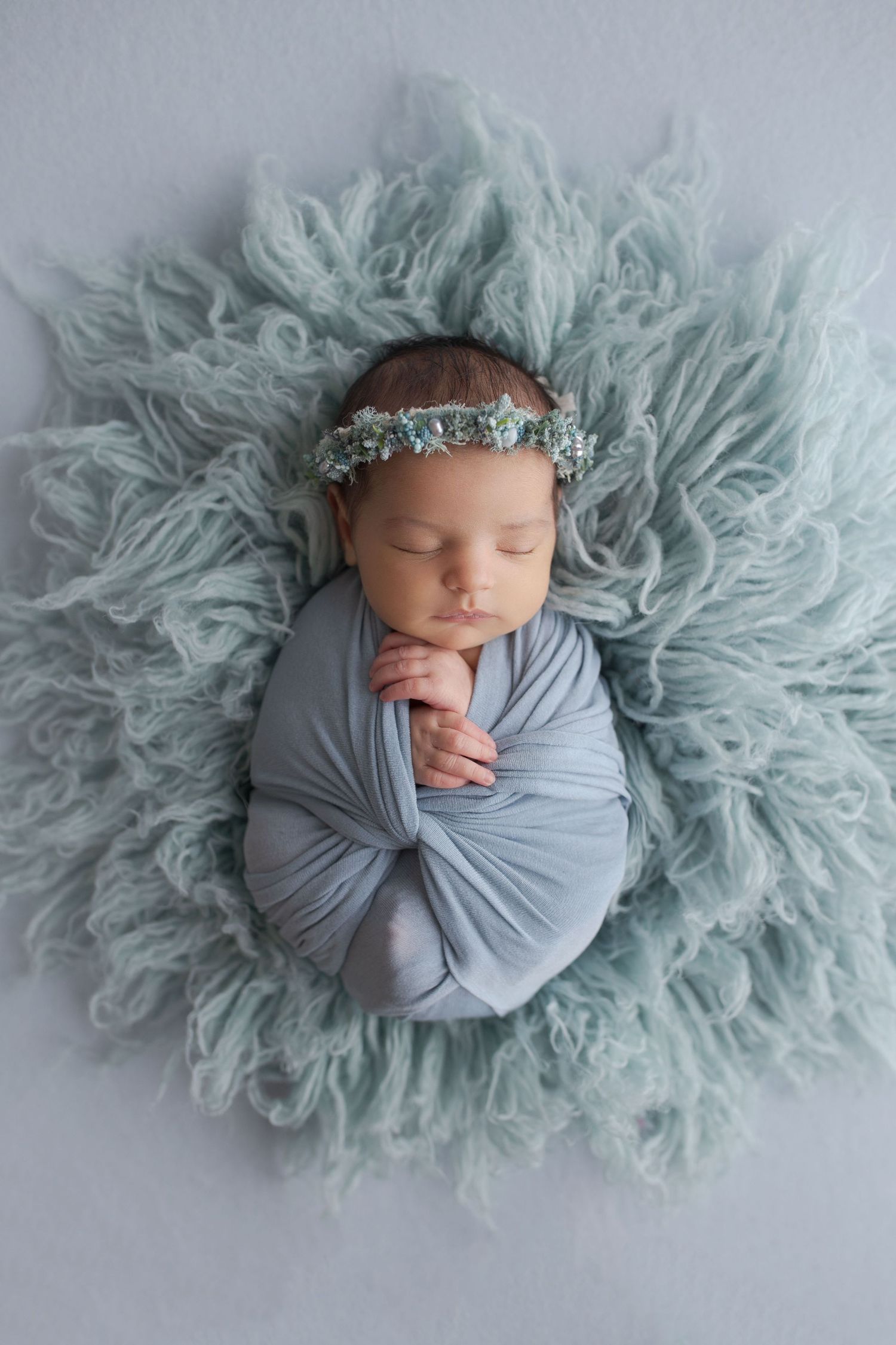 Peaceful newborn baby sleeping on soft blue fur with gray wrap and headpiece.