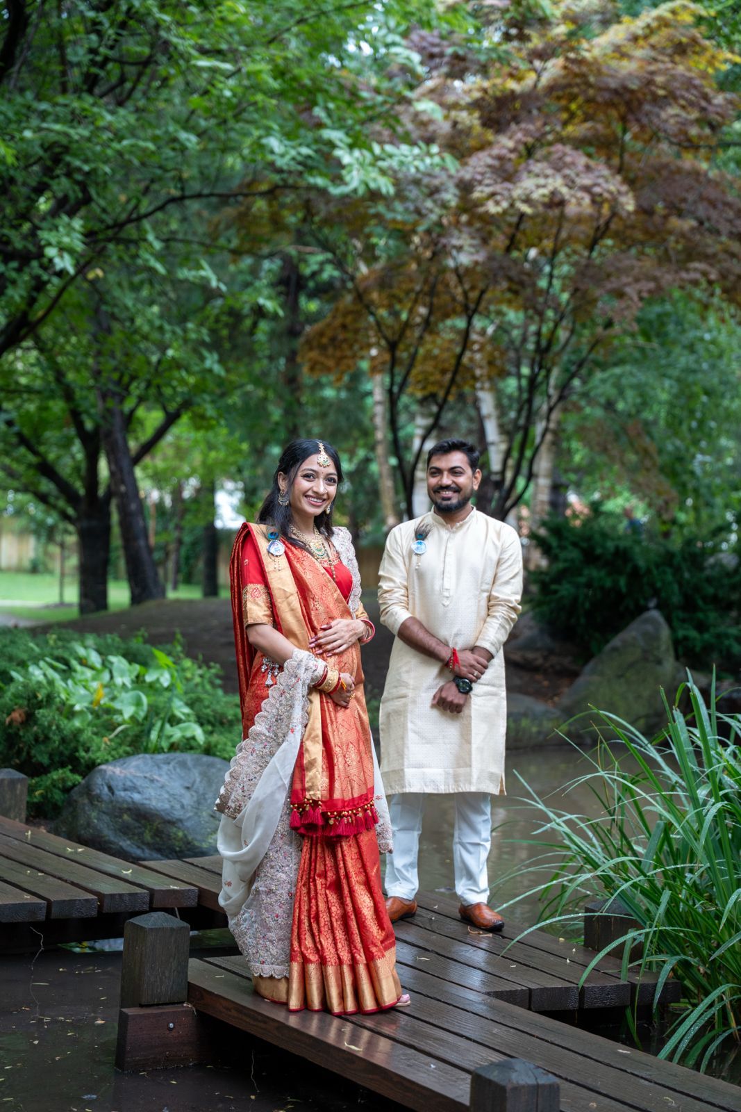 A couple in traditional South Asian attire poses together on a wooden deck surrounded by lush greenery.