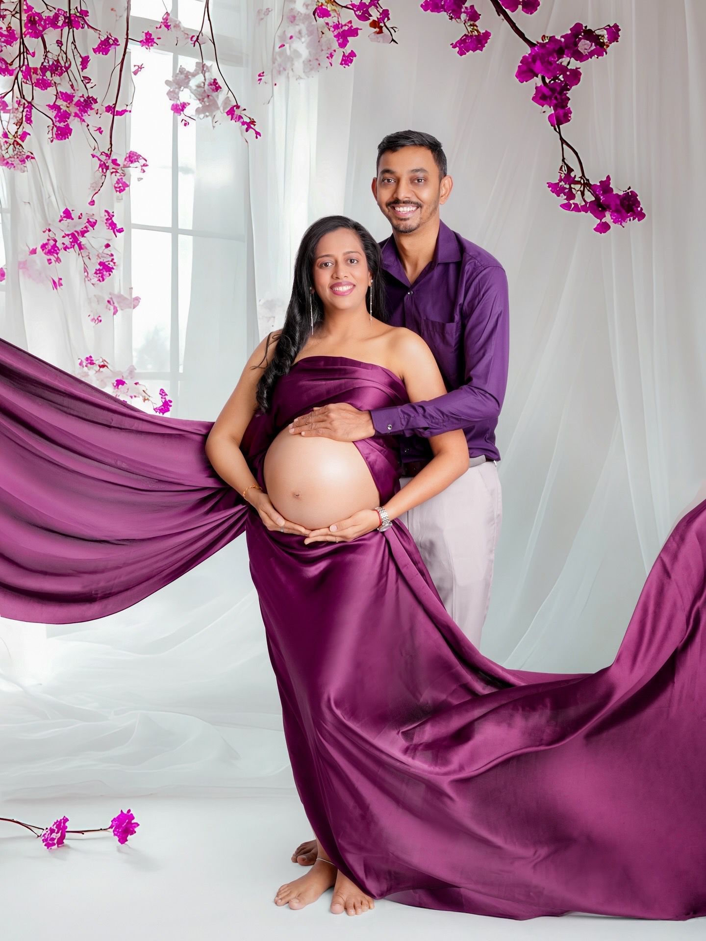 A maternity photoshoot featuring a flowing purple dress and pink orchid flowers in a bright white studio setting.