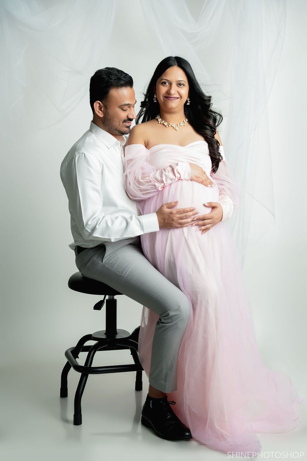 Couple poses for maternity photo with person in pink gown and partner seated on stool against white backdrop.