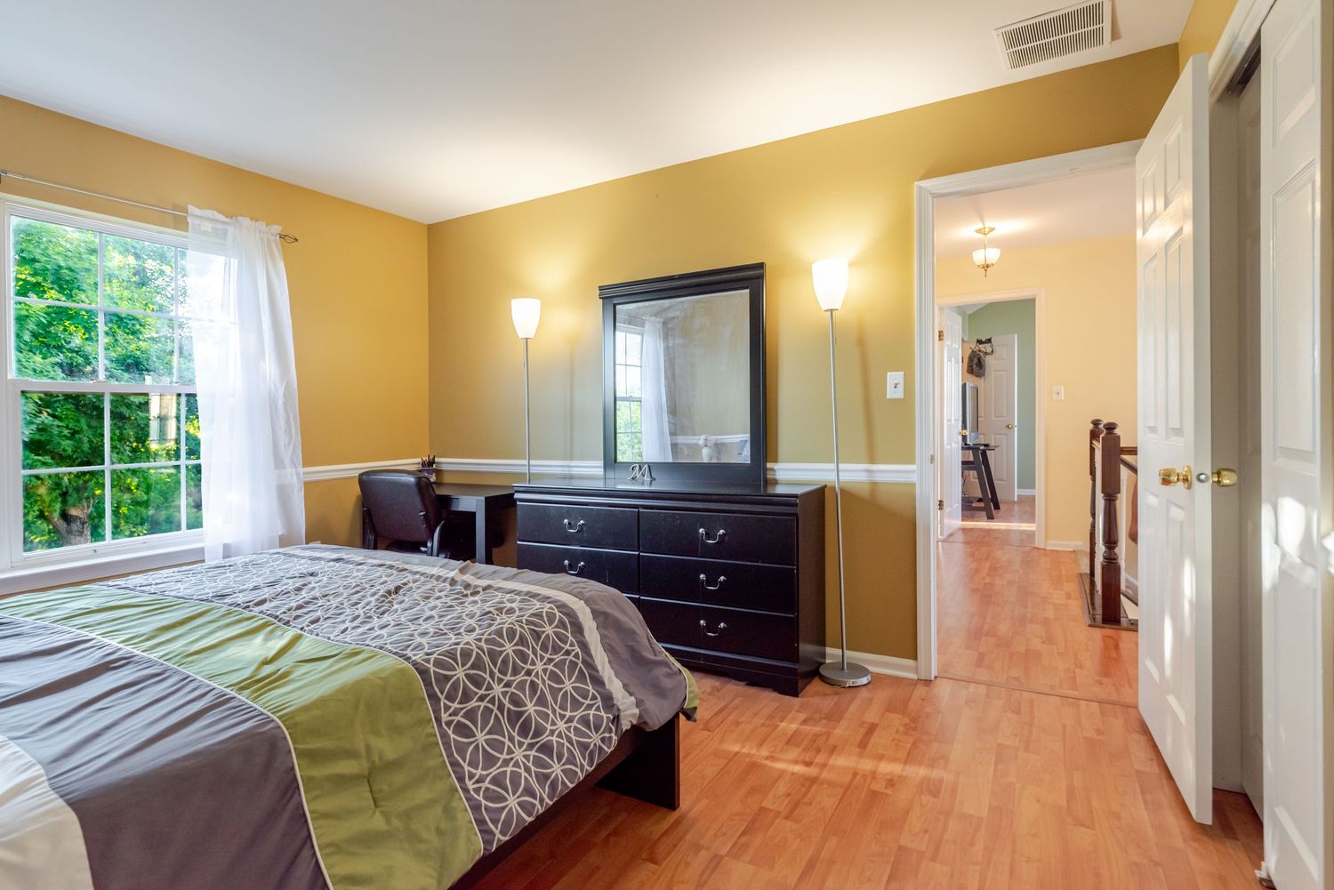 Bright bedroom with hardwood floors, dark wood dresser with mirror, green bedding, and wall sconces in a yellow-painted room.