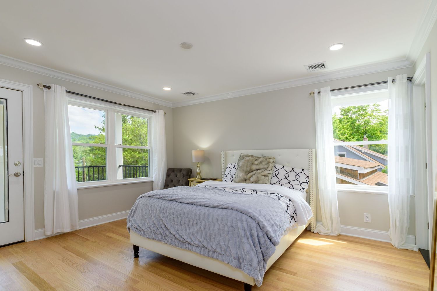 Modern bedroom with light gray walls, hardwood floors, and large windows featuring a gray upholstered bed and white curtains.