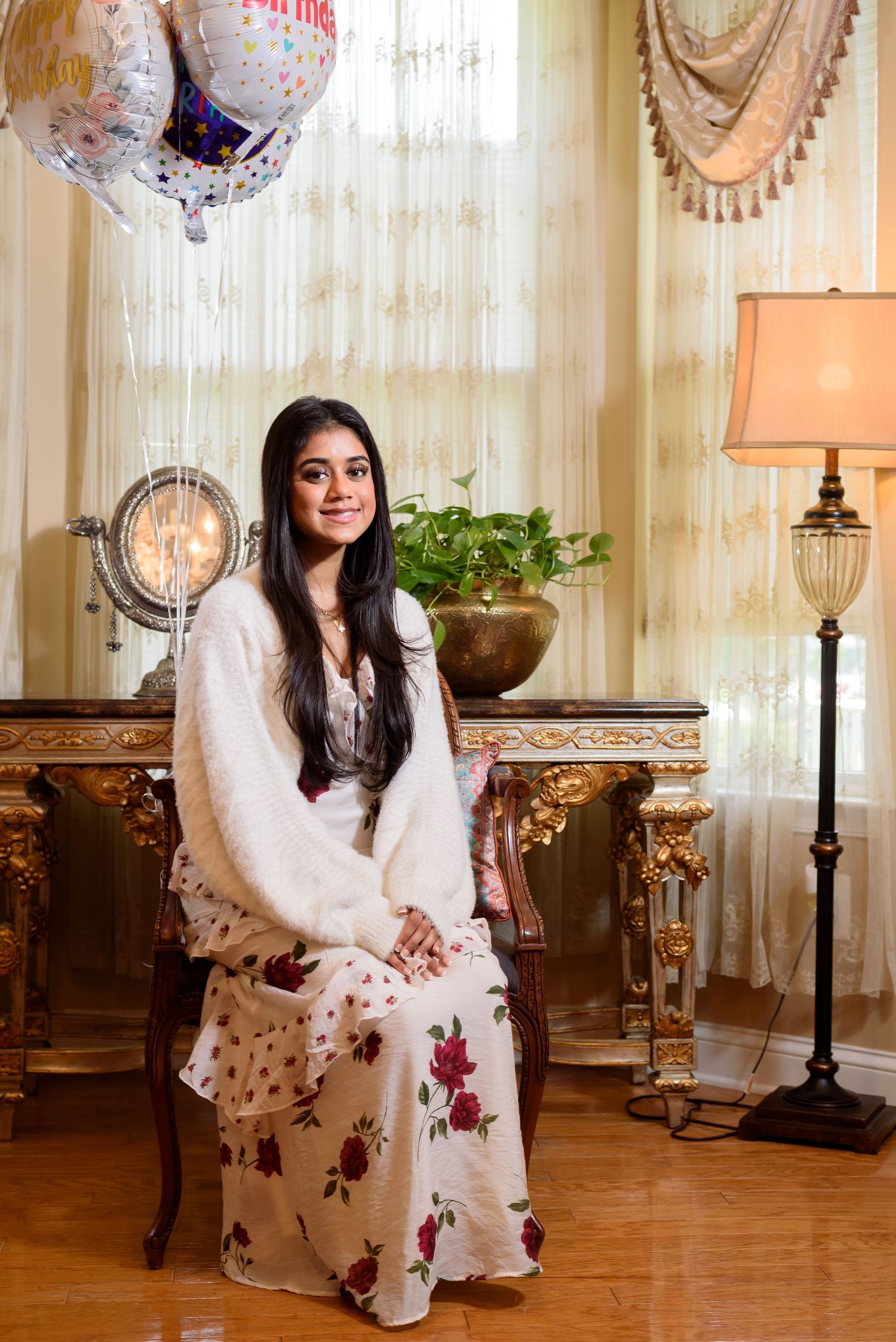 A person in a white floral dress sits elegantly on an ornate chair in a luxurious room with sheer curtains and lamps.
