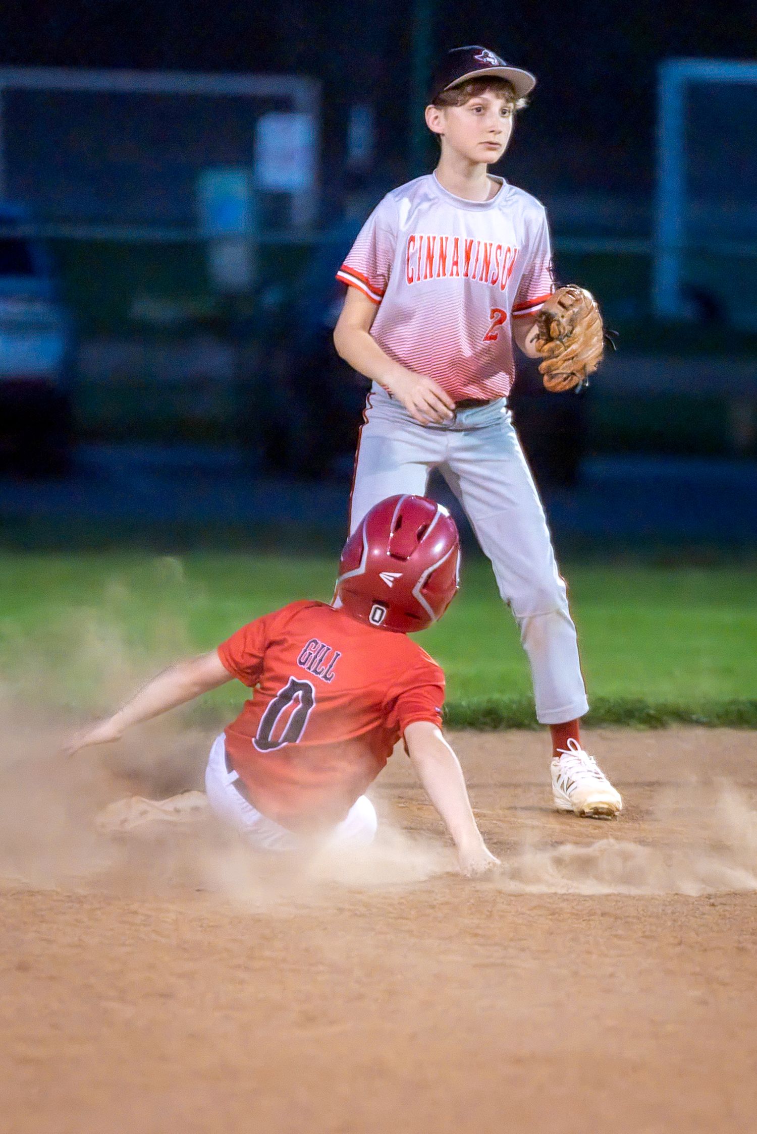 Youth baseball player slides into base as Cinnaminson infielder waits for the ball during a night game.