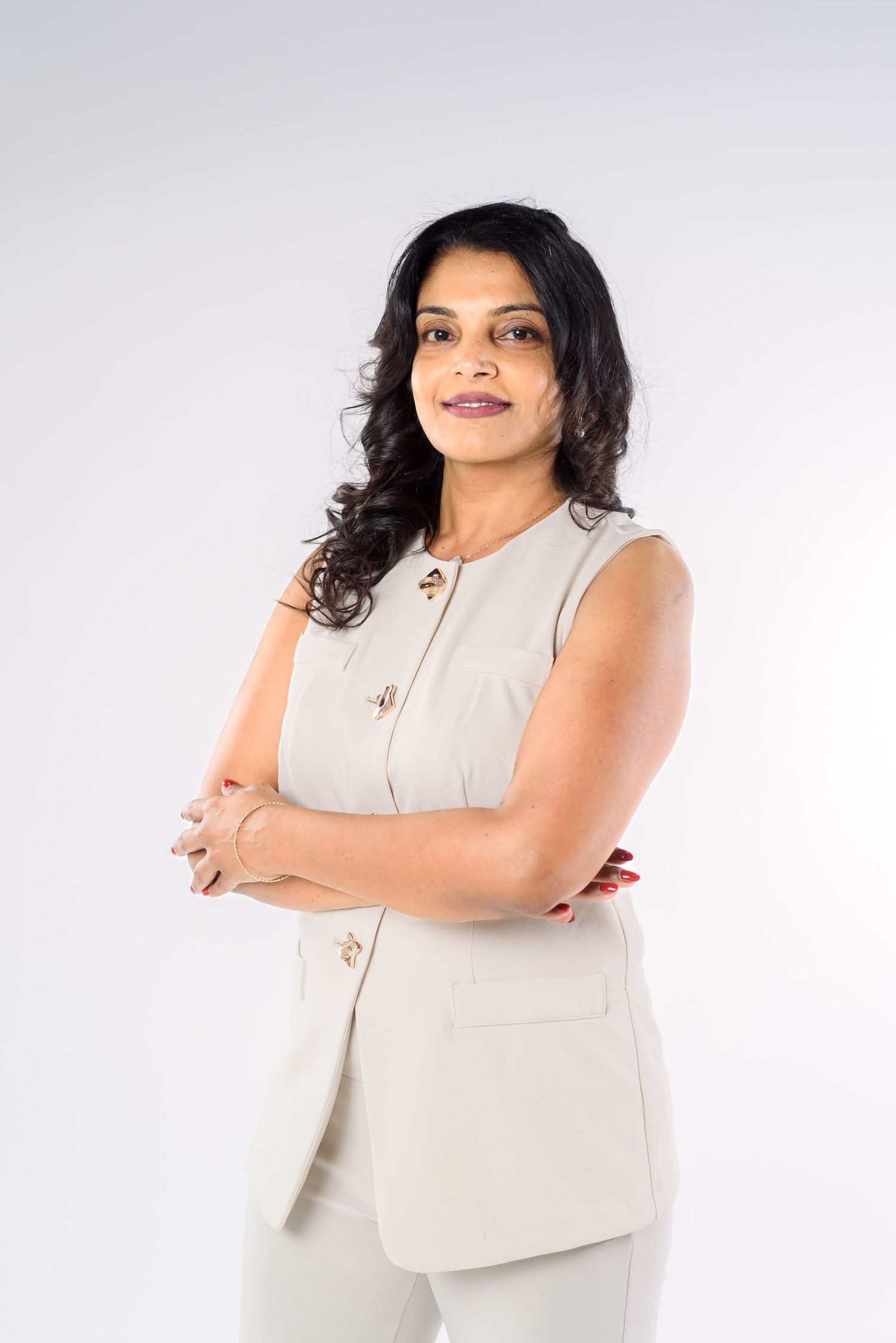 Professional woman in beige sleeveless suit stands confidently with arms crossed against white background.