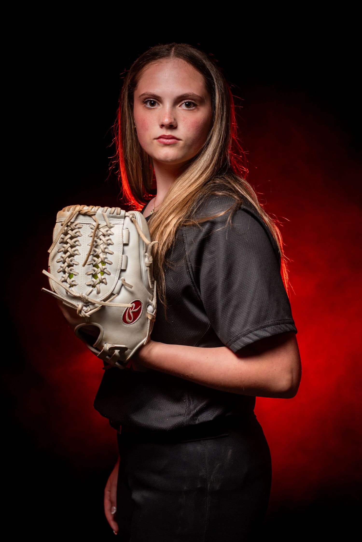 Young female softball player in black uniform holds white fielding glove, posed against dramatic red and black background.