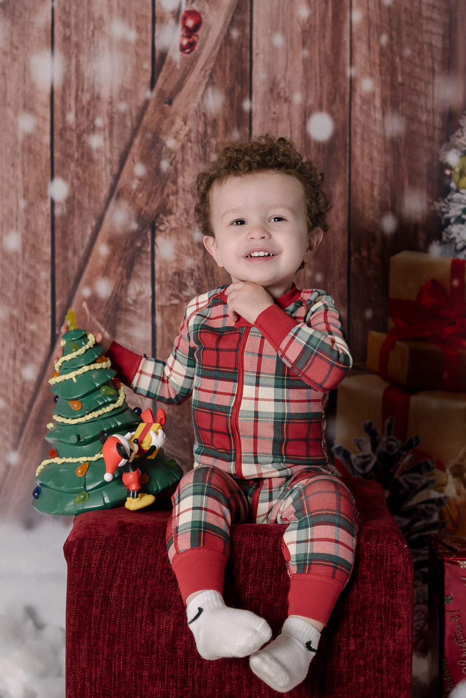 Baby in plaid holiday outfit sits on red couch with decorative Christmas tree during festive photo session.