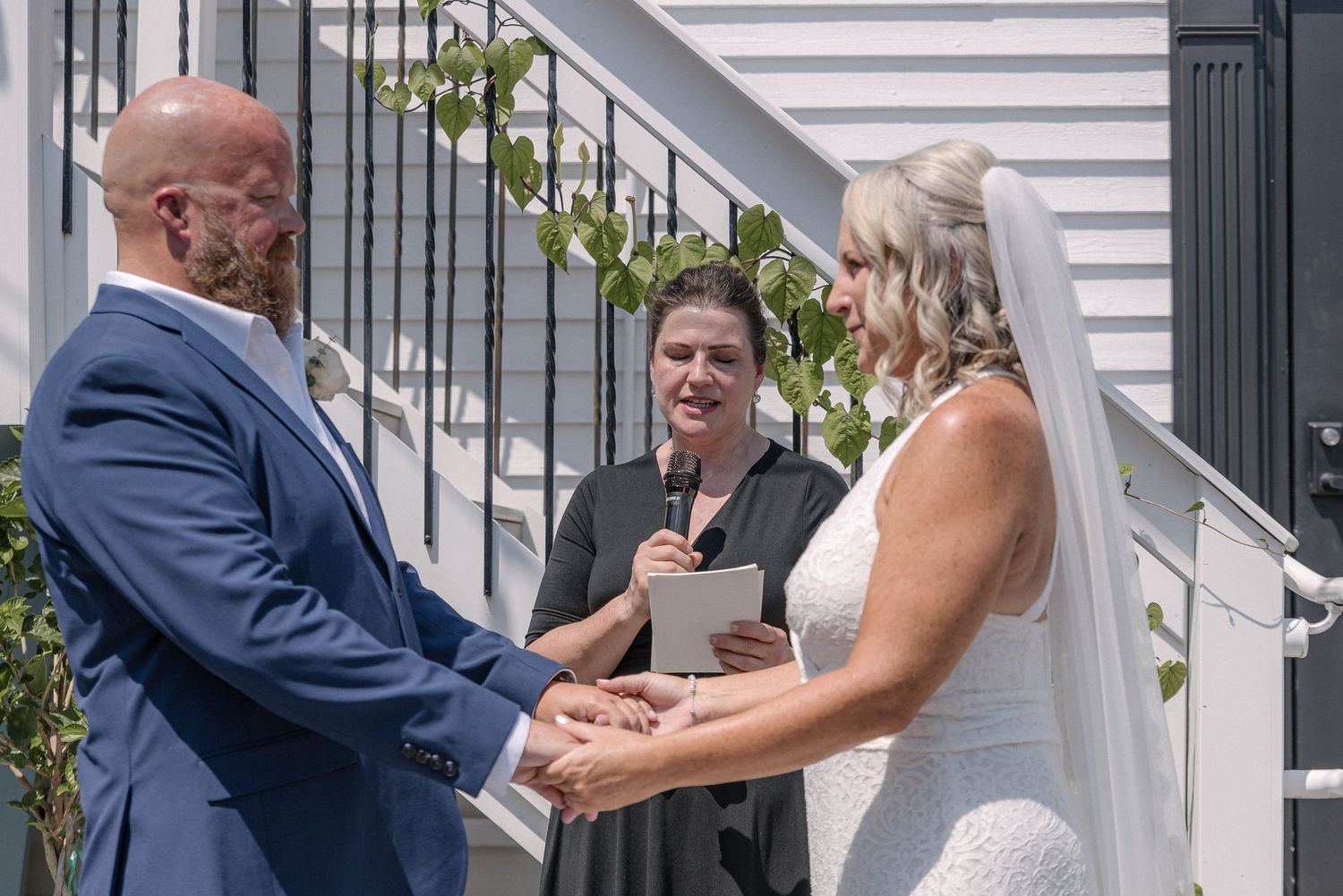 A couple holds hands during an outdoor wedding ceremony with an officiant presiding on a sunny day.