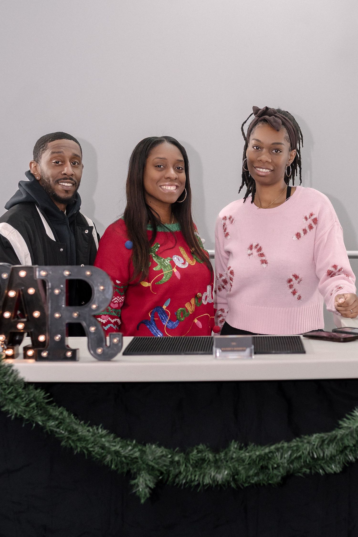 A group of people wearing festive sweaters smile and laugh together at a holiday gathering with decorative garland.