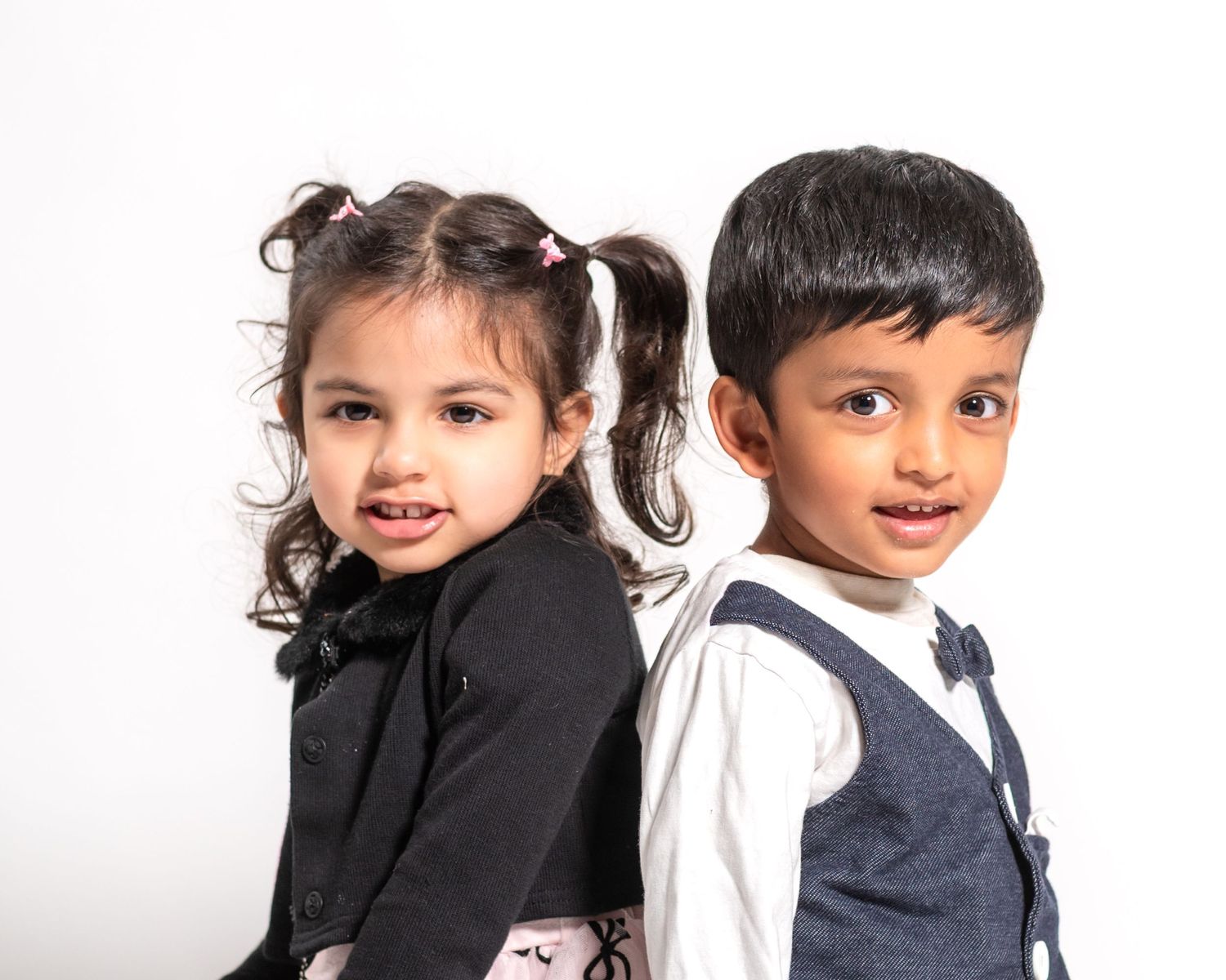 Two young children in formal attire pose back-to-back, smiling against a white background.