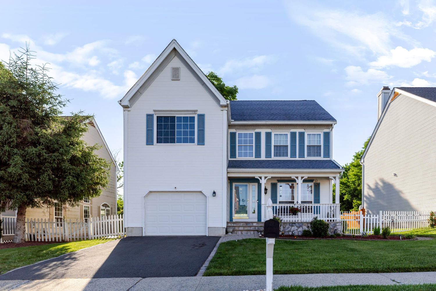 White two-story colonial home with blue shutters, covered porch, attached garage, and well-manicured lawn on a sunny day.