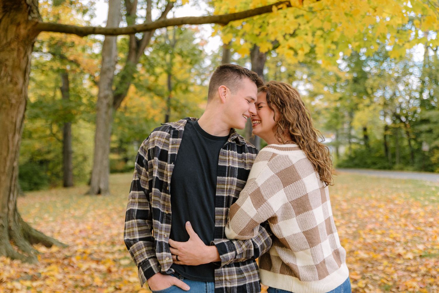 Couple in plaid shirts sharing an intimate moment in a park with golden autumn leaves on the ground.