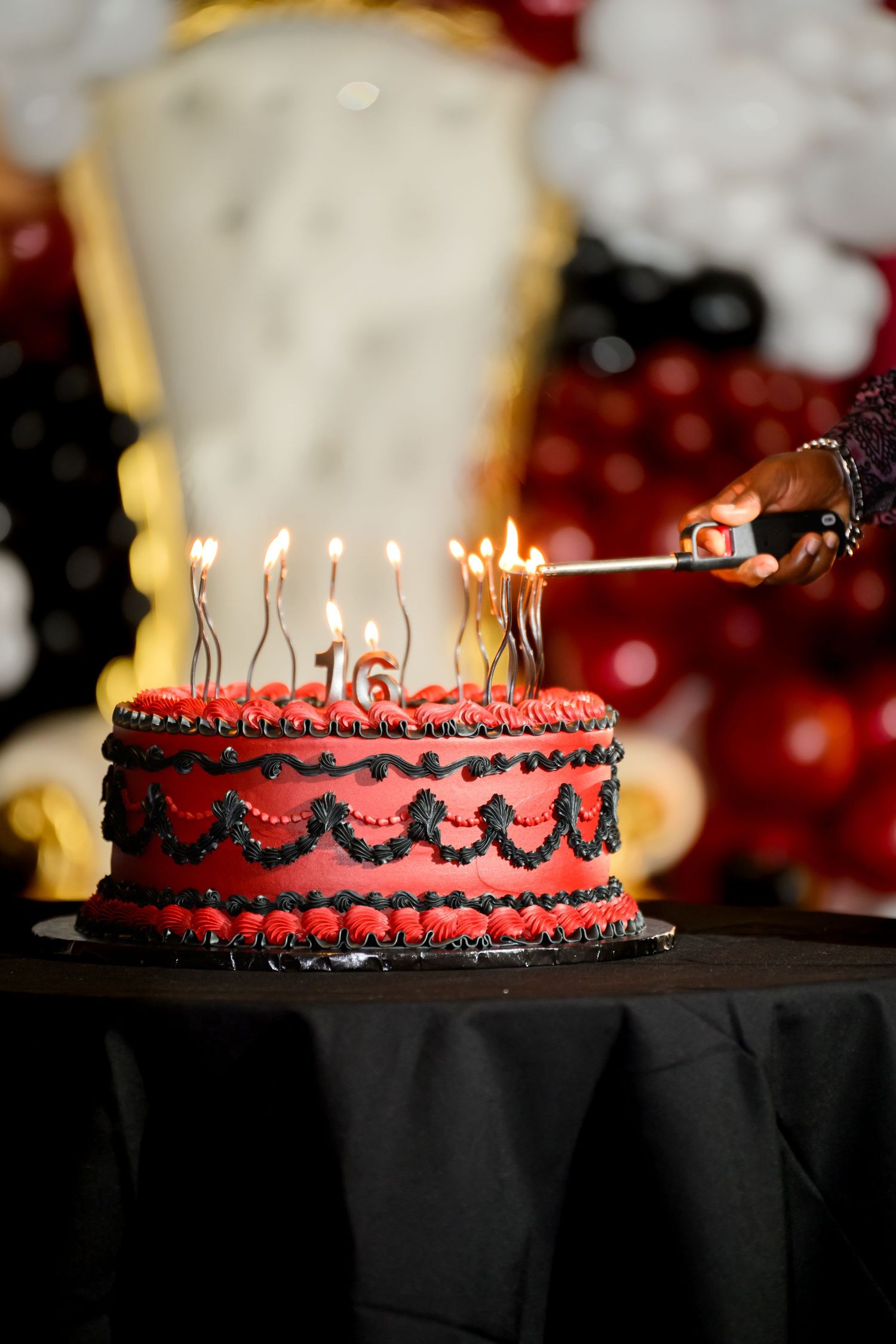 A red and black two-tier sweet 16 birthday cake with lit candles being lit by a lighter.