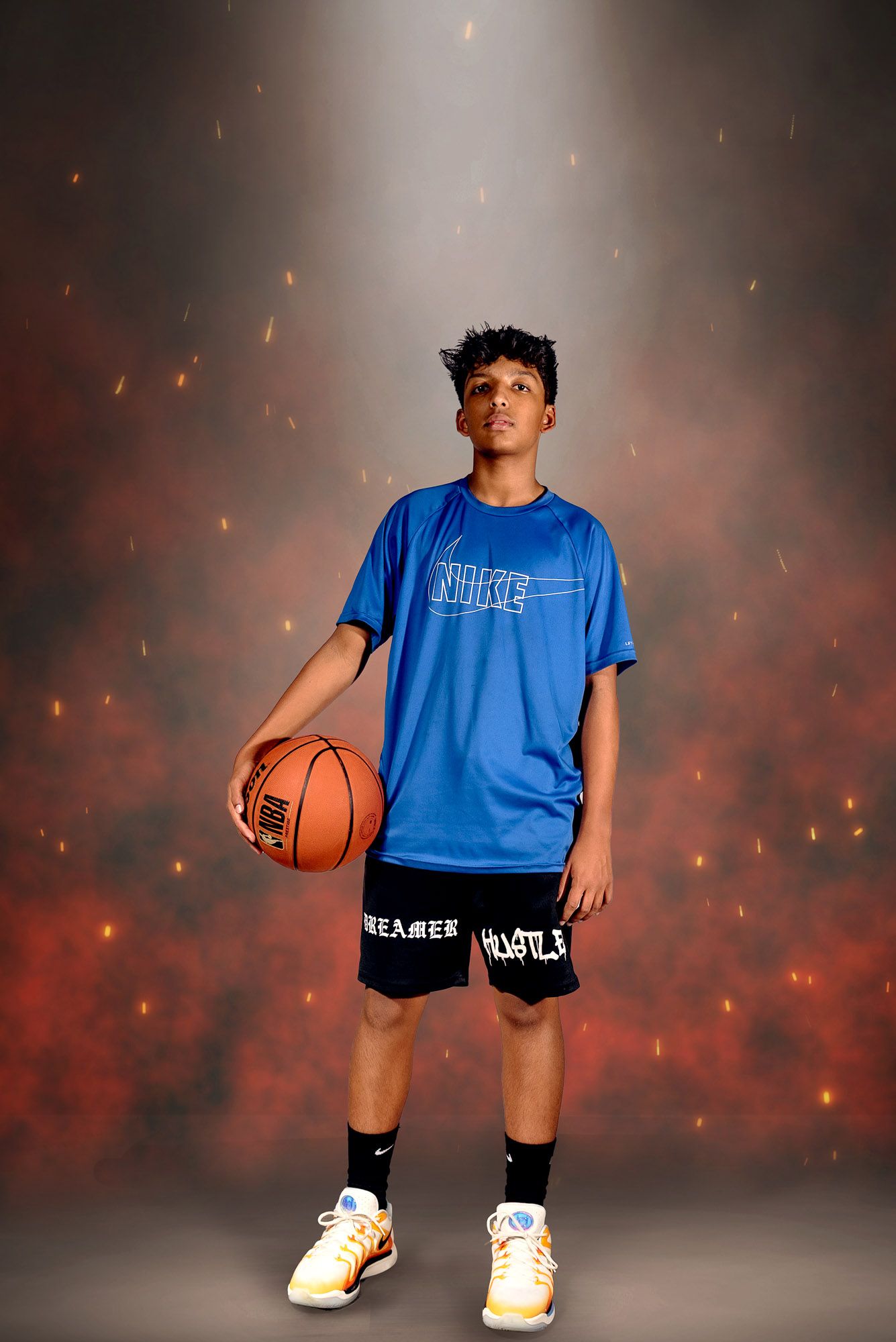 Basketball player in blue jersey and black shorts holds a ball while posing against dramatic background with sparks.