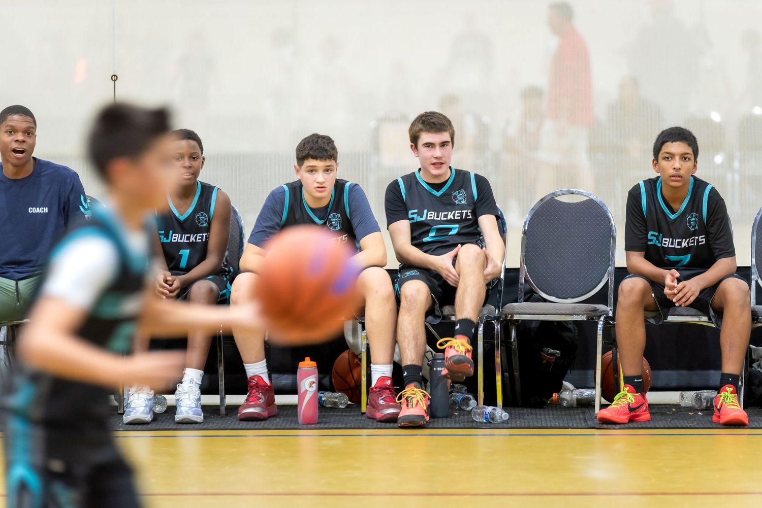 Youth basketball players in teal Rockets jerseys sit on the bench watching a game in an indoor gymnasium.