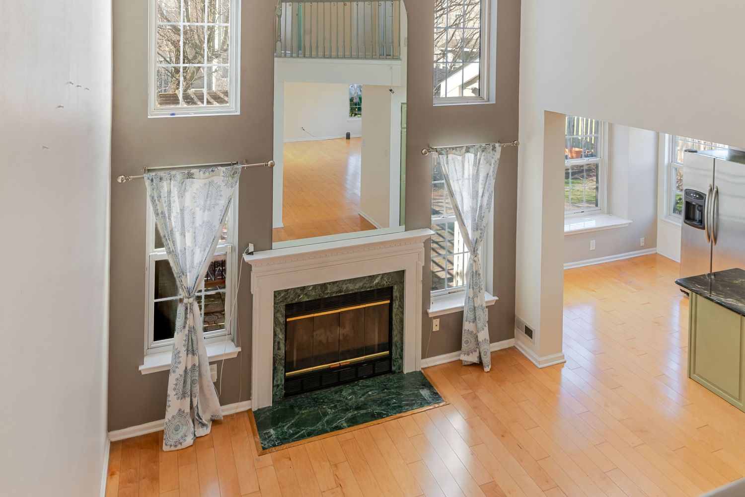 Bright living room with hardwood floors, white fireplace with green marble surround, and tall windows with sheer curtains.